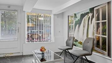 Ein modernes Interieur mit einem Glastisch, zwei grauen Sesseln, einem großen Fenster mit Blick auf ein Auto und Bäume sowie einem Wandgemälde eines Wasserfalls.