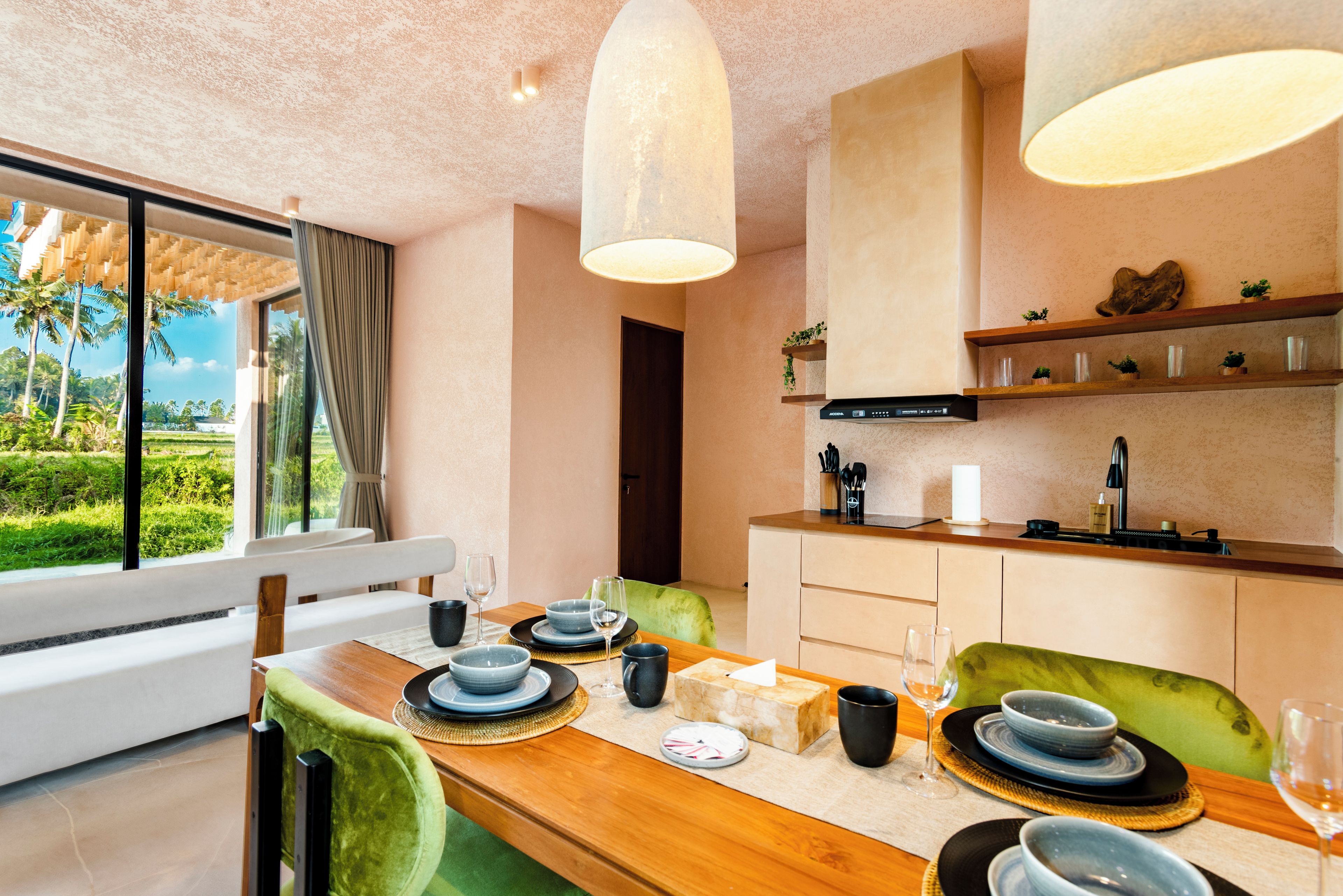 A modern dining area with a wooden table set for two, a kitchen with a built-in stove, and a view of a lush garden through large windows.