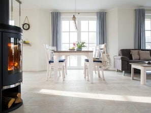 Ein gemütliches Wohnzimmer mit Kamin, Esstisch mit Stühlen, einer Couch und einem großen Fenster mit Vorhängen.