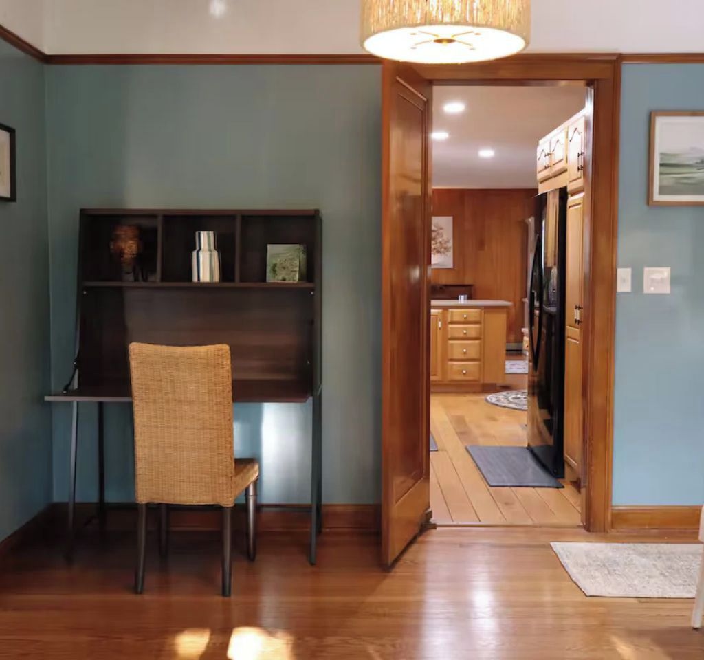 A room with a wooden desk, chair, and a shelf with various items. There is a doorway leading to a kitchen area with wooden cabinets and a refrigerator.