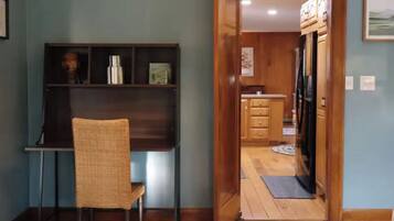 A room with a wooden desk, chair, and a shelf with various items. There is a doorway leading to a kitchen area with wooden cabinets and a refrigerator.