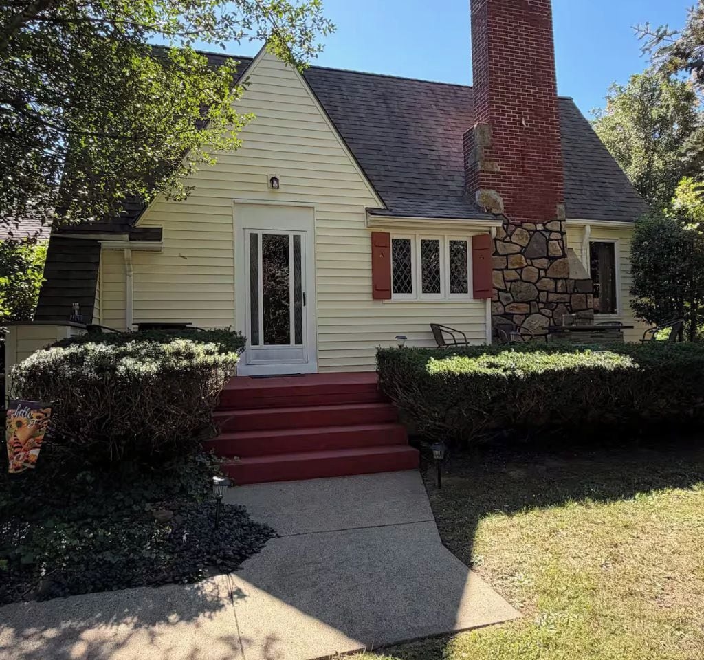 A two-story house with a red front door, a stone chimney, and a small front porch with a bench.