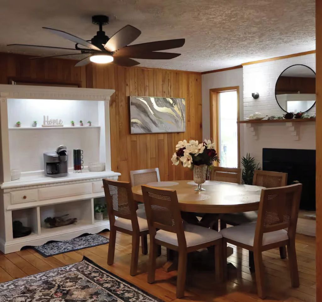 A dining area with a round table, six chairs, a coffee machine, and a wall-mounted shelf with a 'Home' sign.