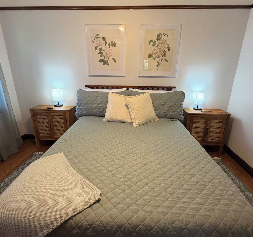 A bed with a quilted cover, two bedside tables with lamps, and two framed botanical prints on the wall.