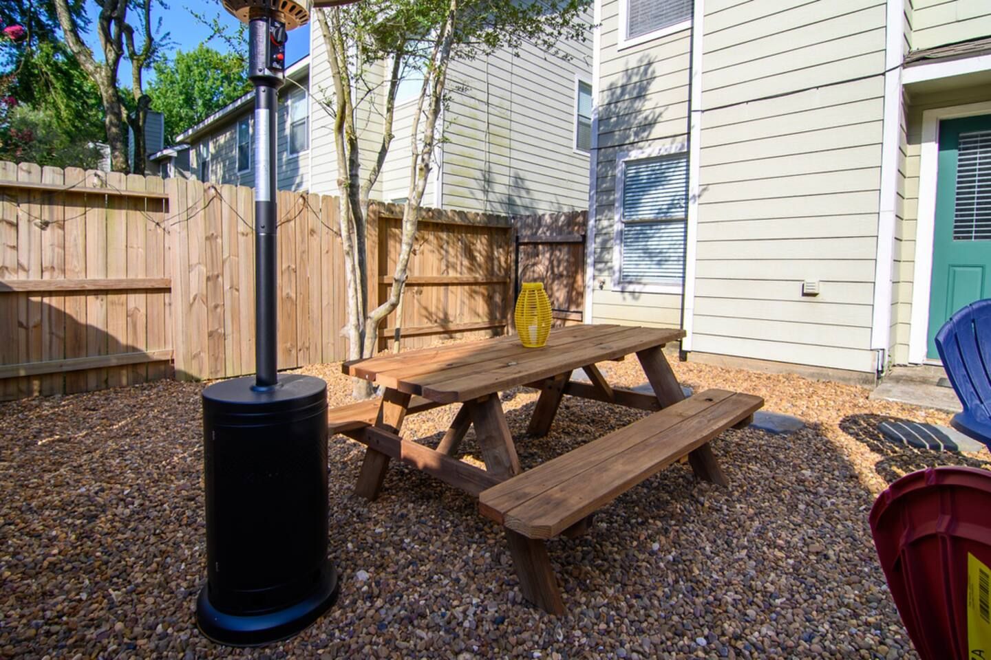 A wooden picnic table with benches, a black grill, and a yellow lantern in a backyard with a wooden fence and a house in the background.