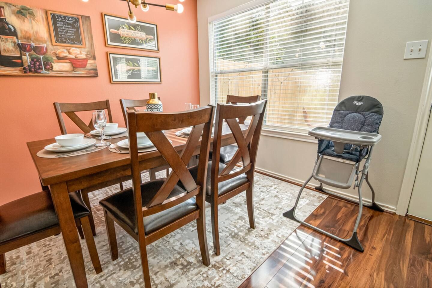 A dining area with a wooden table, chairs, and a high chair. There are framed pictures on the wall and a window with blinds.