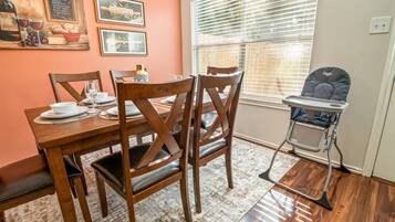 A dining area with a wooden table, chairs, and a high chair. There are framed pictures on the wall and a window with blinds.