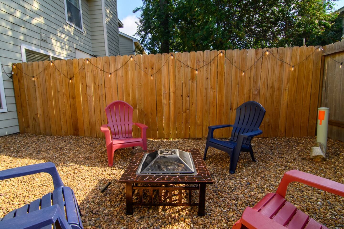 A backyard with a wooden fence, two Adirondack chairs, a fire pit, and string lights.