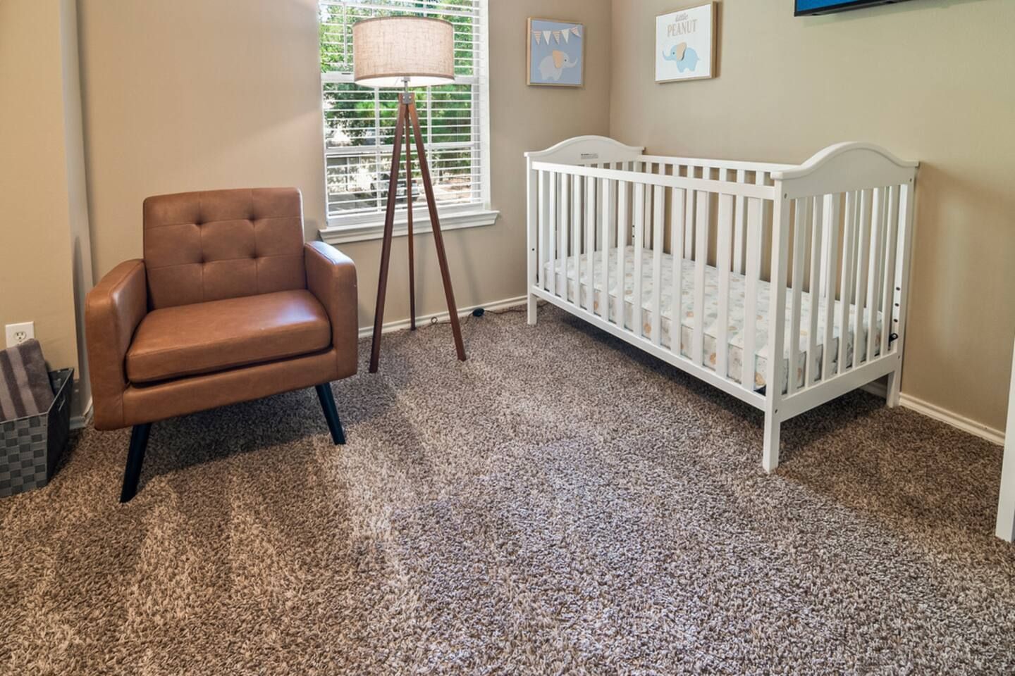 A room with a brown leather chair, a white crib, a floor lamp, and a window with blinds.
