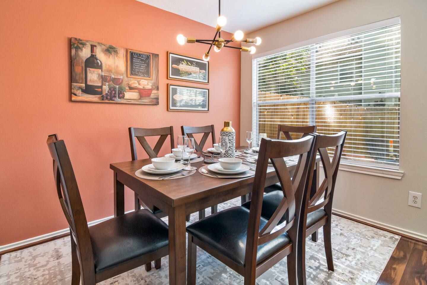 A dining area with a wooden table set for four, surrounded by chairs, with artwork on the walls and a window with blinds.