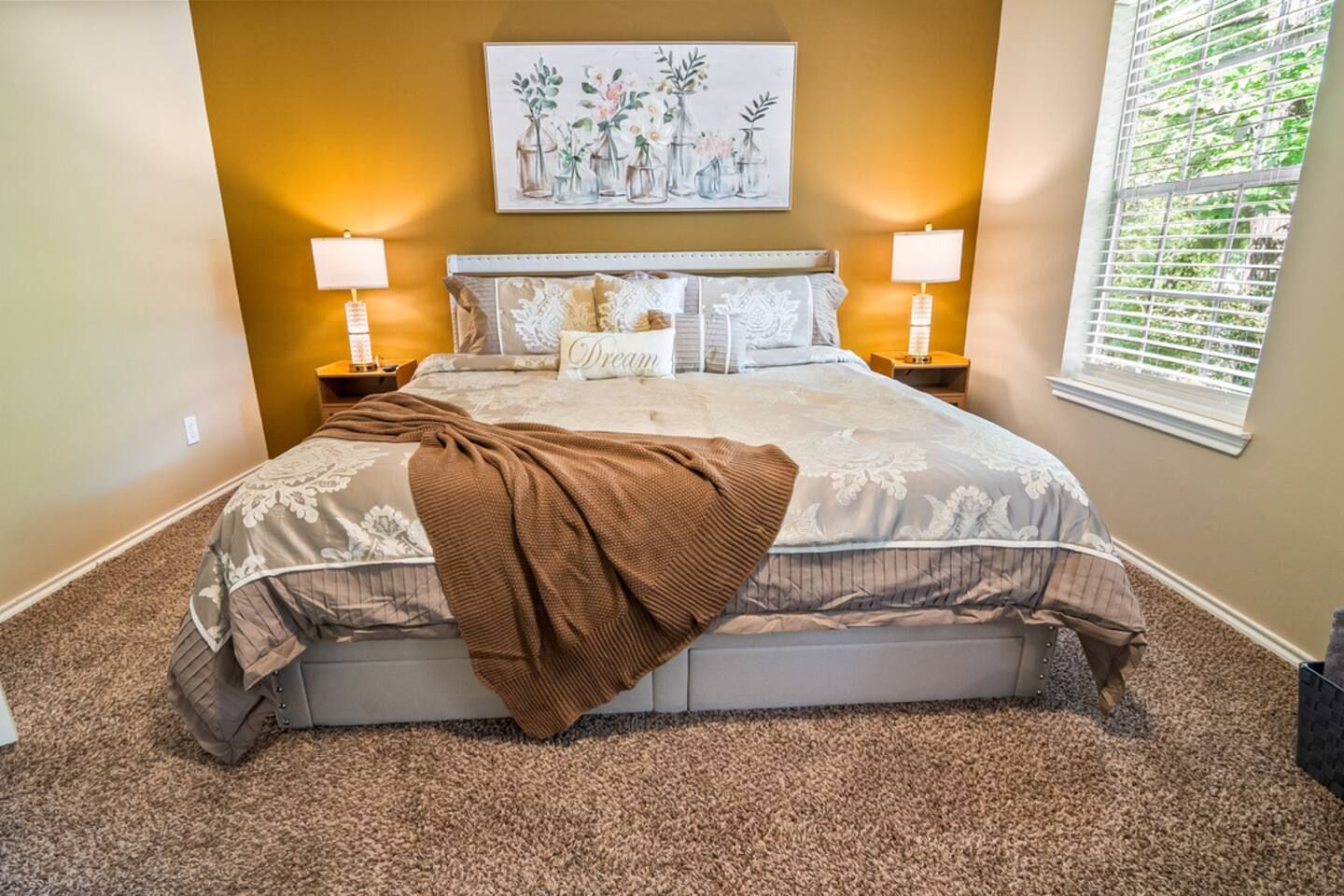 A bedroom with a large bed, two bedside lamps, a wall-mounted painting, and a window with blinds.