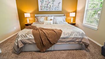 A bedroom with a large bed, two bedside lamps, a wall-mounted painting, and a window with blinds.