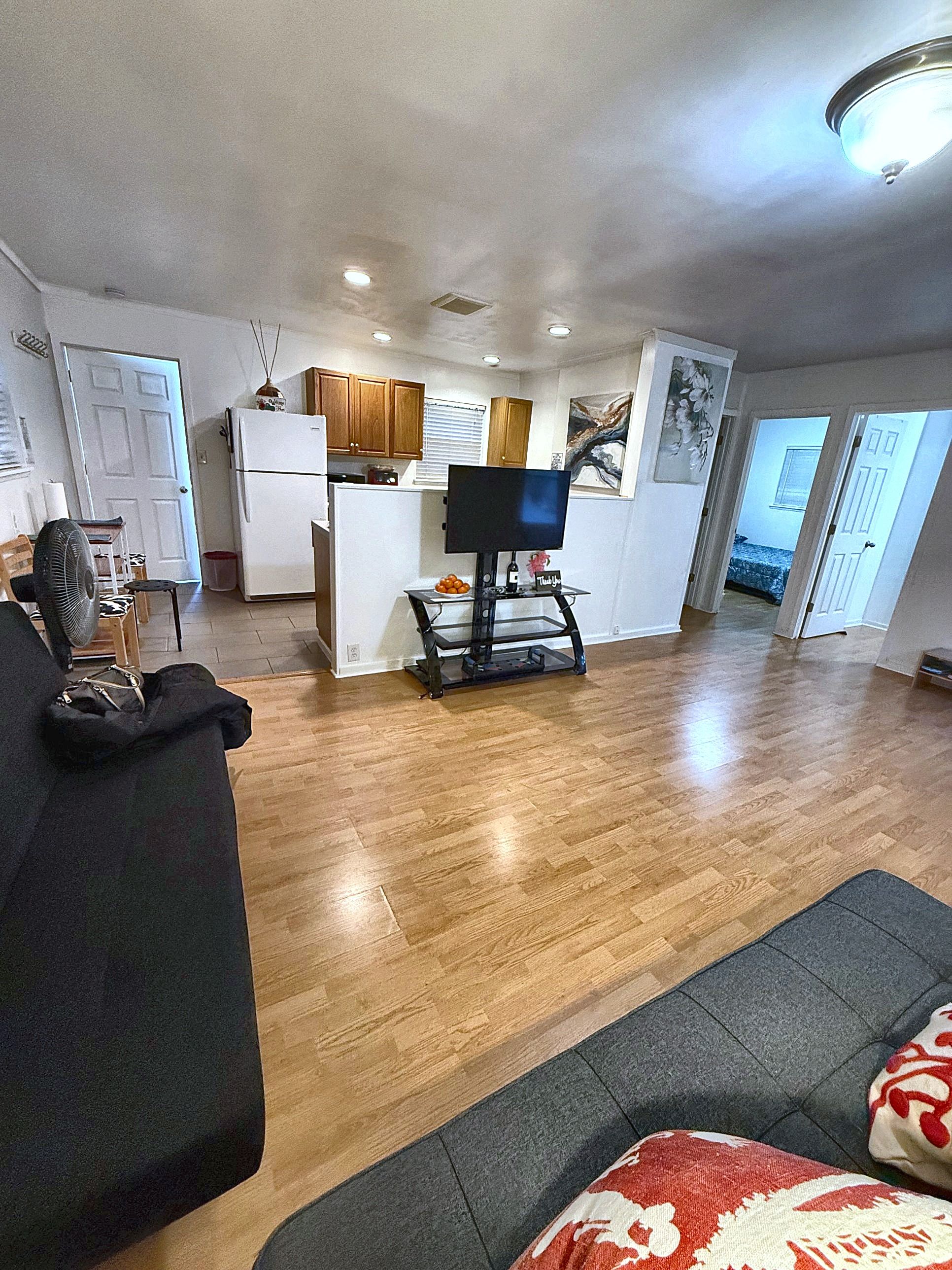 A living room with a black sofa, a television on a stand, wooden flooring, and a kitchen in the background.