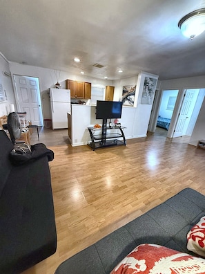 A living room with a black sofa, a television on a stand, wooden flooring, and a kitchen in the background.