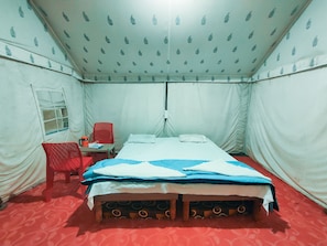 A single bed with a white and blue bedspread, a red chair, a small table with a red chair, and a window with a curtain. - Raj Resort (Dholavira)