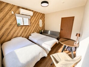 Habitació amb dos llits, paret de fusta, finestra, aire condicionat, mirall, porta i taula rodona amb una làmpada.
