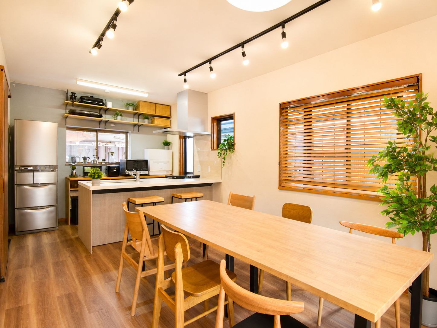 A modern kitchen with a dining area, wooden furniture, and a large window with blinds.