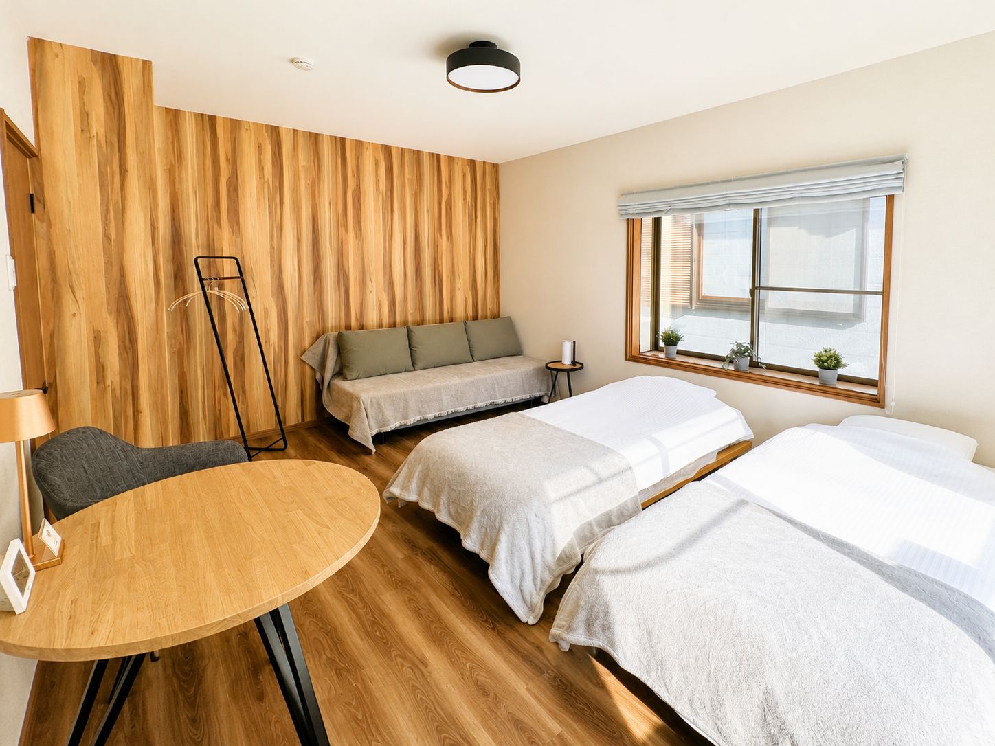 A hotel room with a wooden wall, a round table, two beds, a sofa, and a window with blinds.