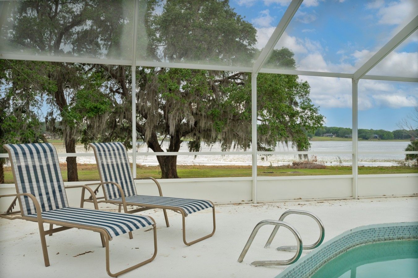Ein überdachter Außenbereich mit zwei gestreiften Liegestühlen, einem Pool und Blick auf Strand und Bäume.