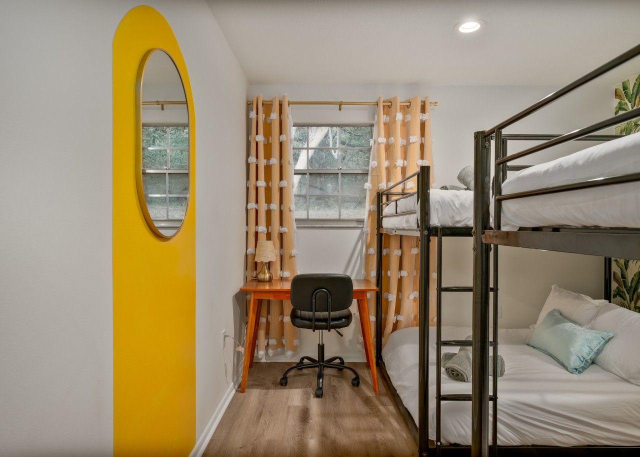 A compact room with a bunk bed, a desk with a chair, a mirror, and a window with curtains.