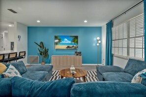 Een woonkamer met een blauwe wand, een flatscreentelevisie, een eethoek met stoelen en een grote plant.