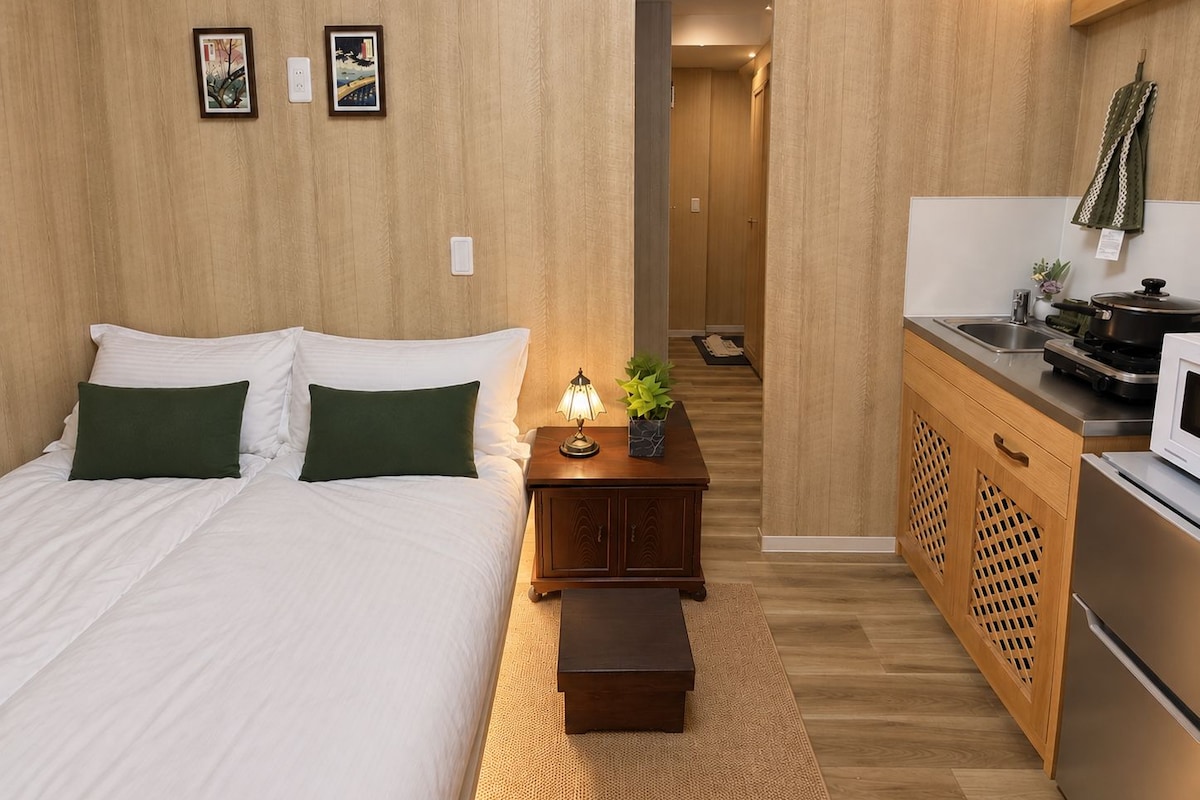 A hotel room with a bed, bedside table with a lamp, a small plant, and a kitchenette with a microwave.