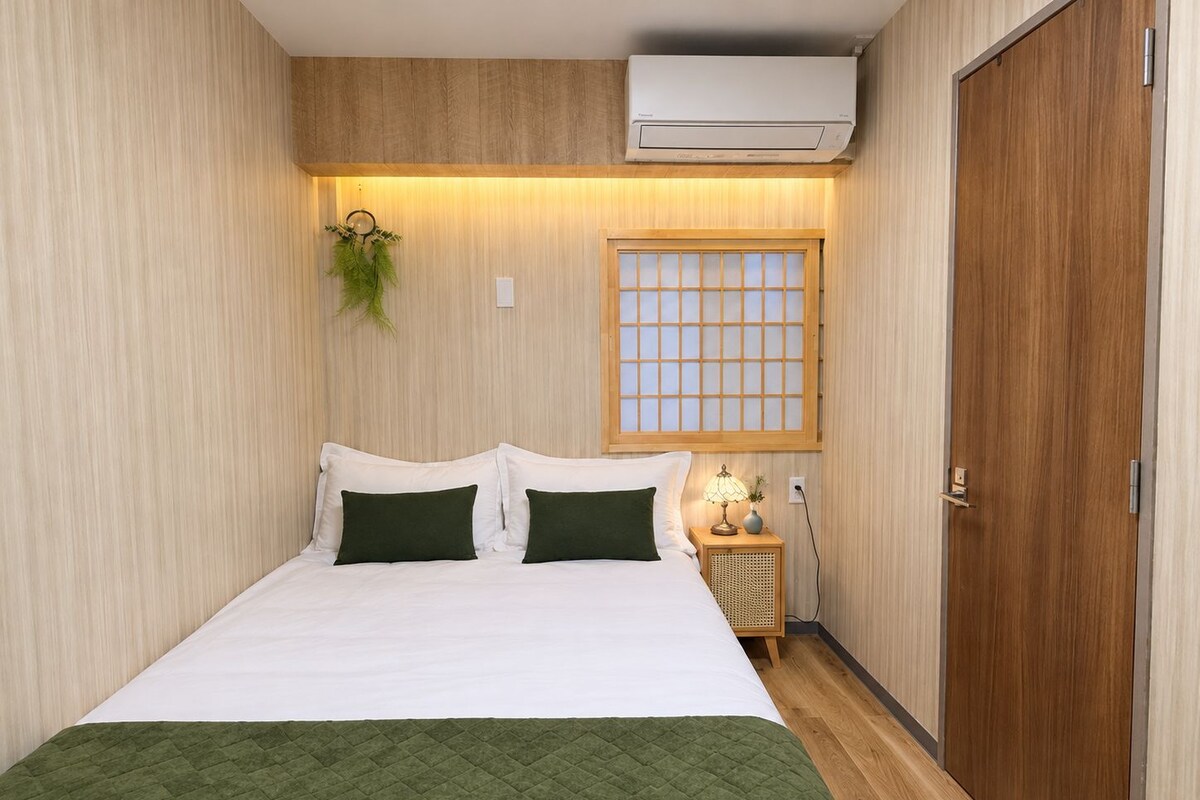 A hotel room with a bed, a window with a grid pattern, an air conditioning unit, a bedside lamp, and a door.