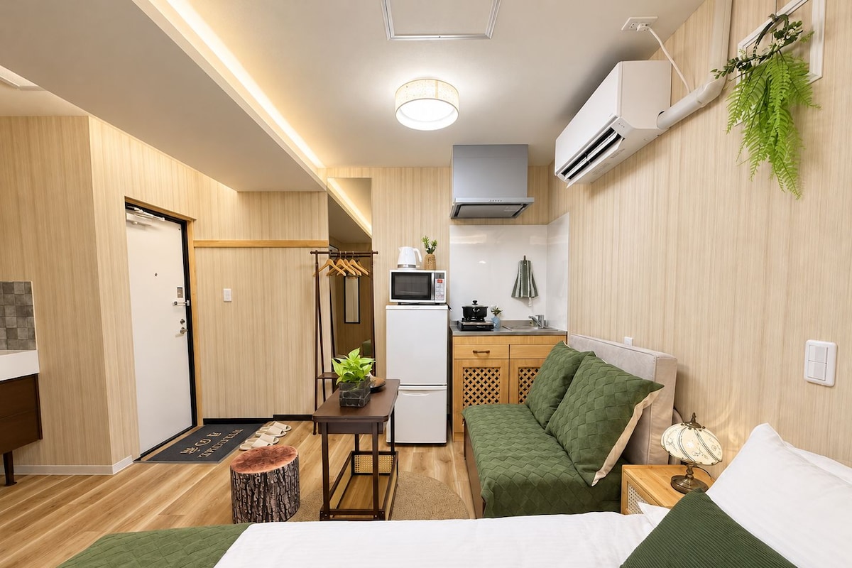 A compact hotel room with a bed, a small desk, a kitchenette, and a seating area.