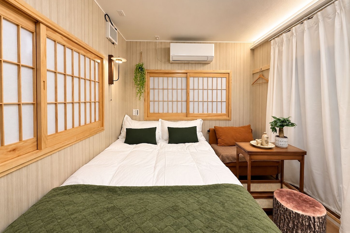 A room with a bed, a small table, a hanging plant, and a window with a view.