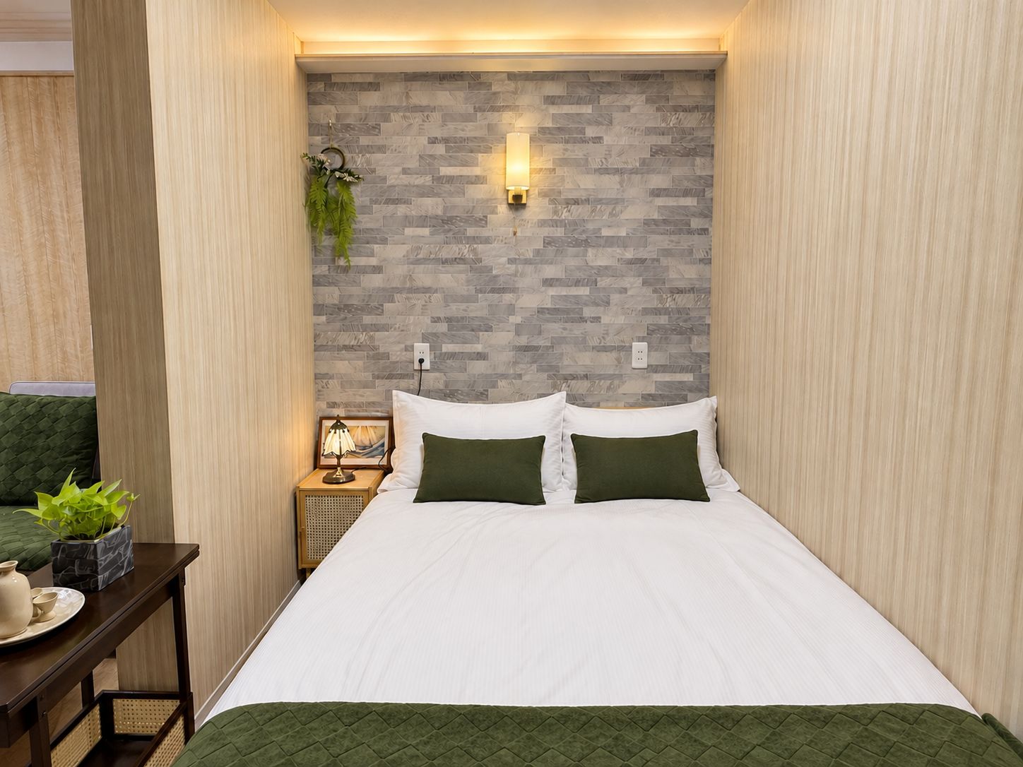 Een netjes opgemaakt bed met witte beddengoed en groene kussens, een nachtkastje met een lamp, en een wandlamp.