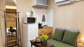 Een compacte kitchenette met een magnetron, fornuis en wastafel. Er staat een klein tafeltje met een plant en een plank met hangende spullen. Een groene, beklede bank staat tegen de muur.