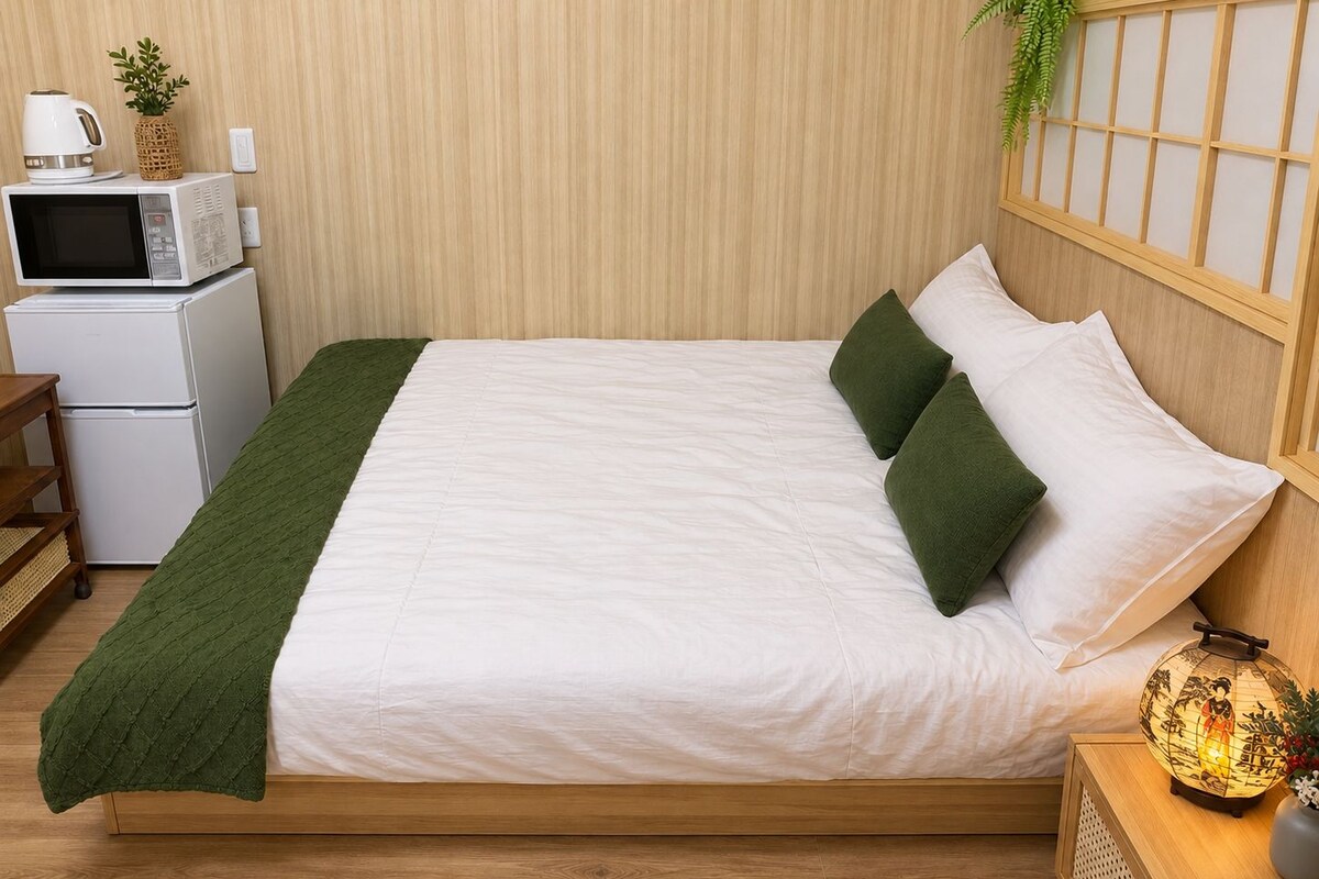 A neatly made bed with a green throw, a bedside lamp, and a small table with a plant.