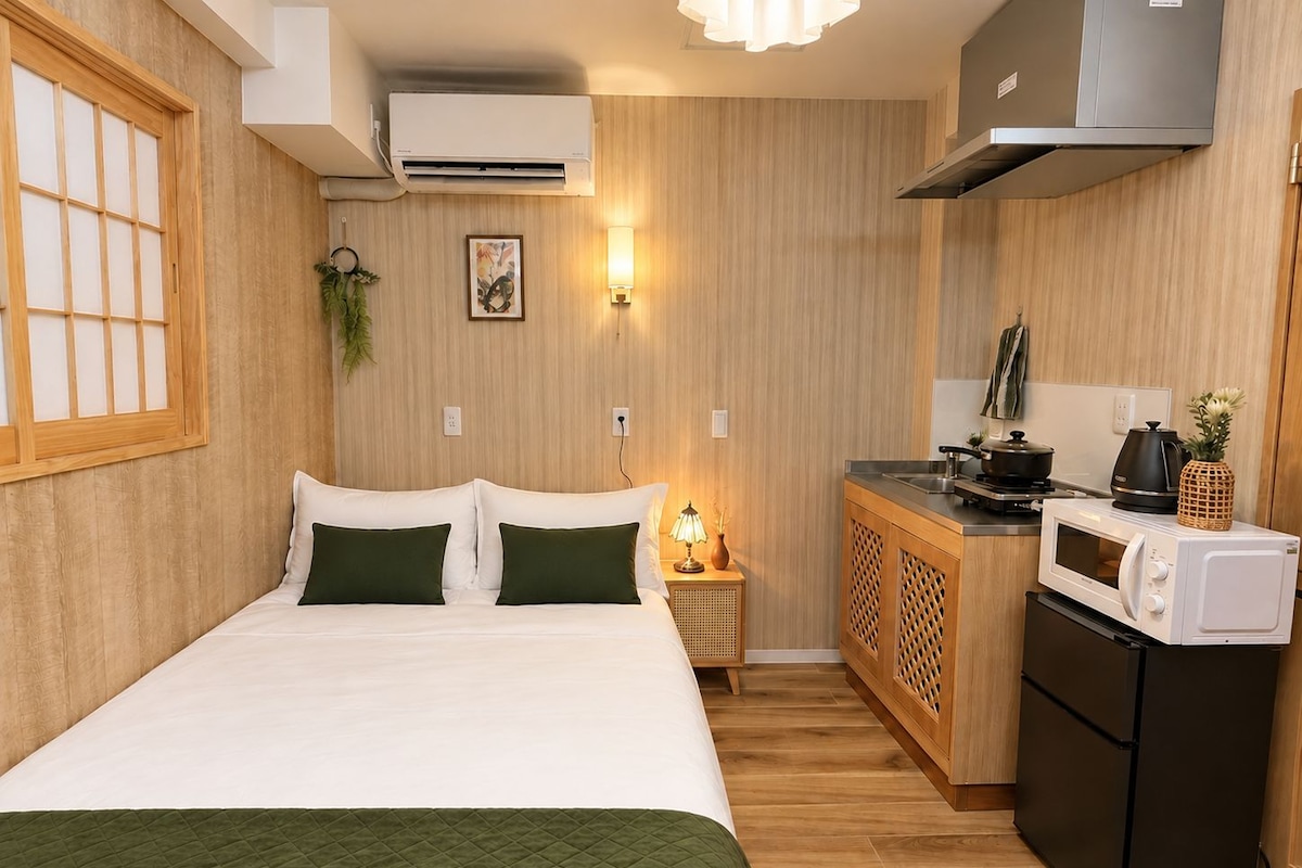 A compact hotel room with a bed, bedside table, lamp, and kitchenette including a microwave and refrigerator.