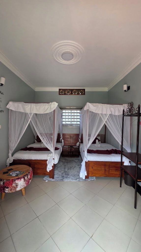 A room with two beds, each with a canopy, a small round table, and a window with blinds.