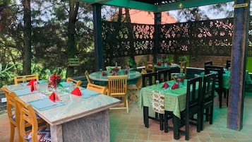 An outdoor dining area with tables and chairs, a pergola structure, and a view of trees.