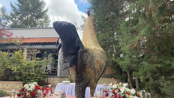 Un área de comedor al aire libre con mesas dispuestas para una comida, rodeada de árboles y una gran escultura de un ave.