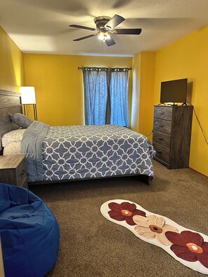 A bedroom with a bed, a dresser, a desk, a chair, a ceiling fan, and a window with curtains.