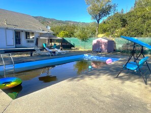 Ein Pool mit Liegestühlen und einer überdachten Liegefläche, umgeben von einem Zaun und Bäumen.