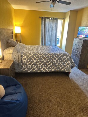 Ein Schlafzimmer mit Bett, Nachttisch, Lampe, Kommode, Deckenventilator und Fernseher.