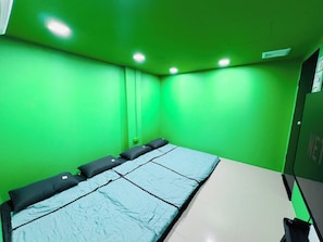 Une pièce aux murs verts, avec un lit simple doté d’un matelas bleu et une petite table accompagnée d’une chaise.