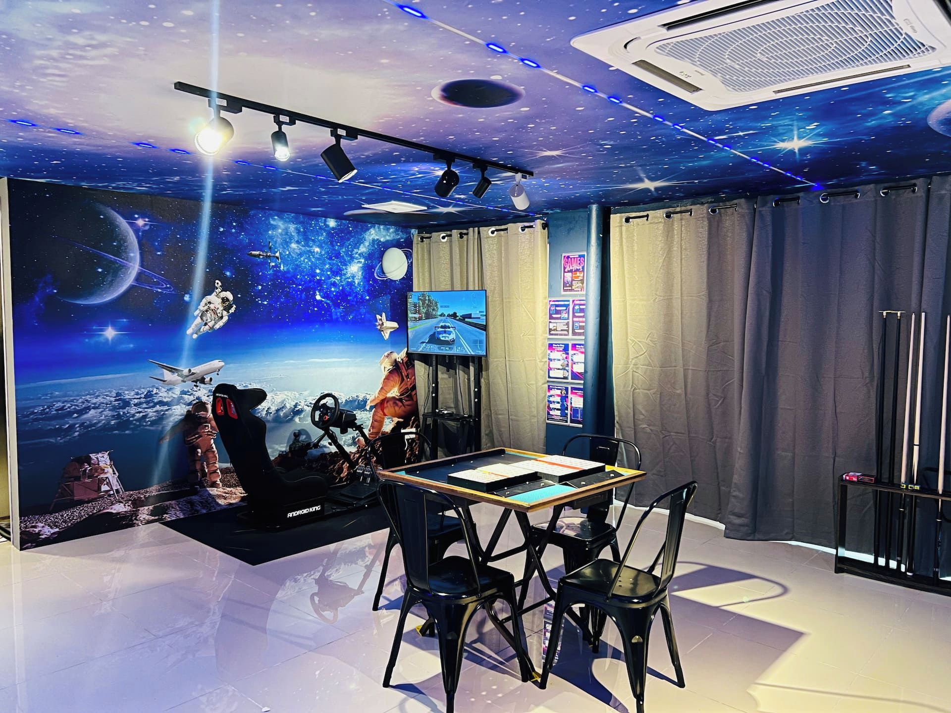 A room with a space-themed wall mural, a mounted television, a table with chairs, and a ceiling fan.