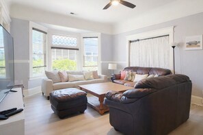 Ein Wohnzimmer mit Sofa, Hocker und Couchtisch, großen Fenstern mit Jalousien, einem Deckenventilator und einer Lampe.