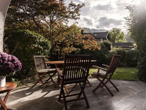 An outdoor patio with a wooden table and chairs, surrounded by lush greenery and a view of a house in the background. - Holiday Apartment With 1 Bedroom (Garmisch-Partenkirchen)