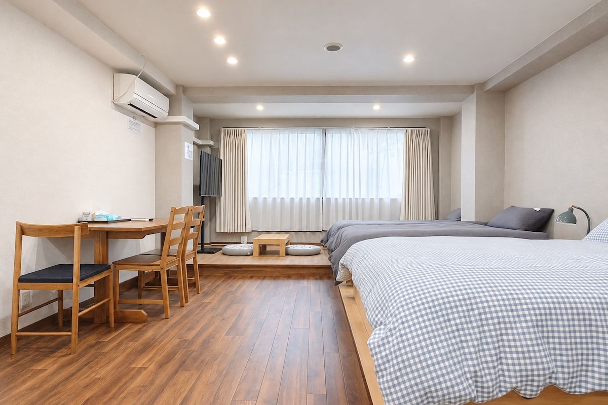 A hotel room with a wooden floor, a desk with chairs, a bed with a checkered bedspread, and a window with curtains.