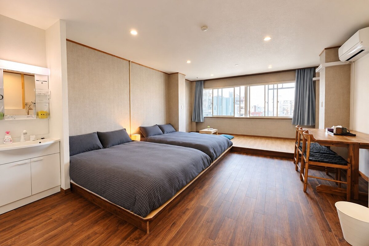 A hotel room with a wooden floor, two beds with grey bedding, a desk with chairs, a small table, and a window with curtains.