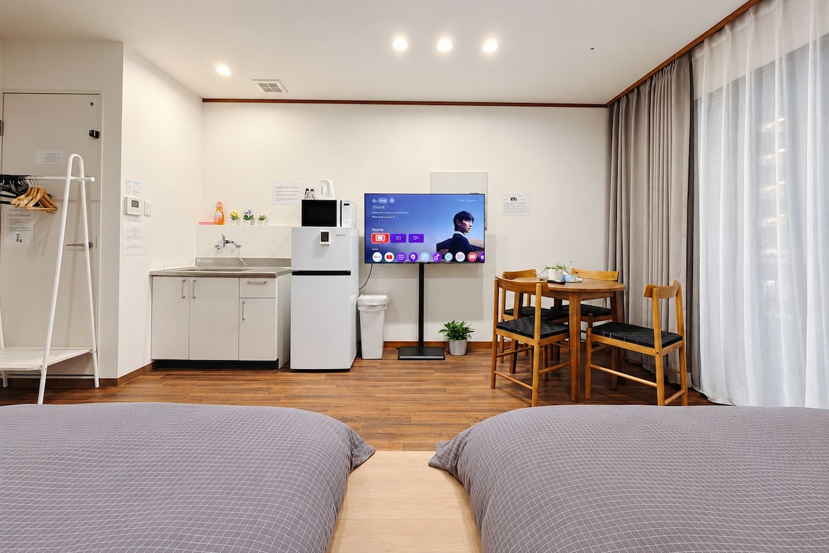 A modern hotel room with a kitchenette, a flat-screen TV, a dining area with a table and chairs, and two beds.