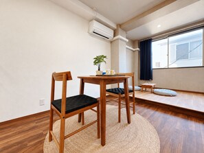 A room with a wooden dining table, two chairs, a small side table, a potted plant, and a window with curtains. - Sanmi Tsutenkaku (Osaka)