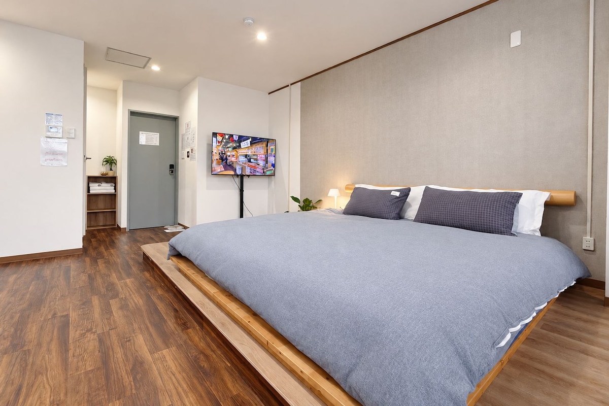 A modern hotel room with a large bed, wooden flooring, a flat-screen TV, and a small desk area.