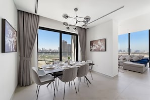 A modern dining area with a table set for four, large windows offering a city view, and a contemporary chandelier. - FIRST CLASS | 3BR | Elevated Luxury Near Downtown (Sobha Hartland)