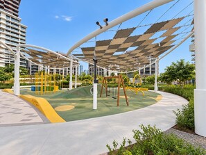 An outdoor play area with swings and climbing structures, surrounded by a white canopy and greenery. - FIRST CLASS | 1BR | Elegant Escape At City Walk (City Walk)